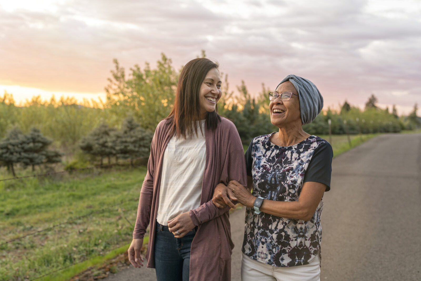 A senior mixed race woman with cancer walks with her adult daughter at sunset down a rural road. They are relaxing and staying active together. The affectionate pair are talking and walking with arms linked.