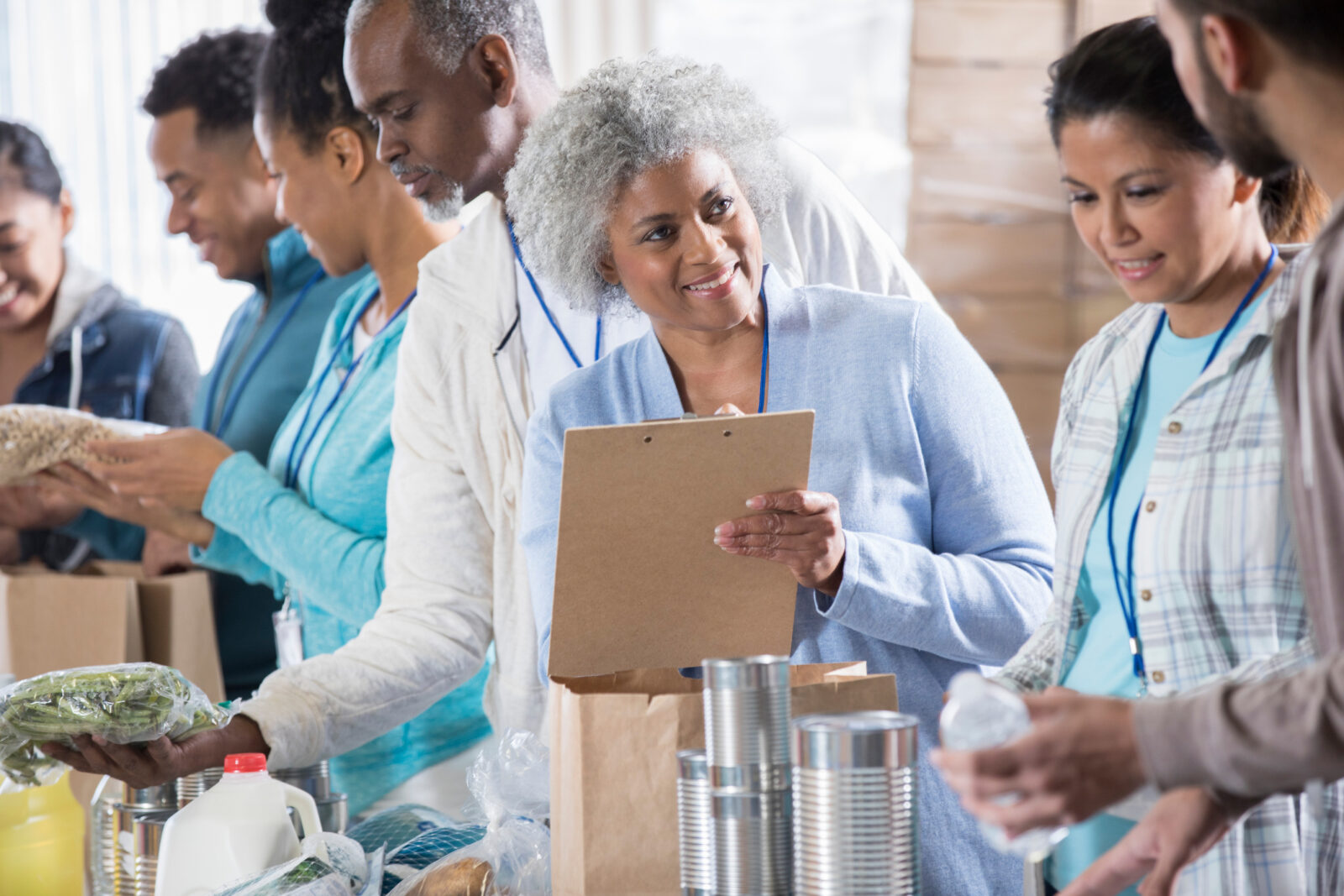 A cheerful Black senior woman holds a clipboard and discusses inventory with two food bank volunteers.